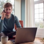 Un homme dans la trentaine en recherche d’emploi, assis à un bureau avec un ordinateur portable et un téléphone à la main