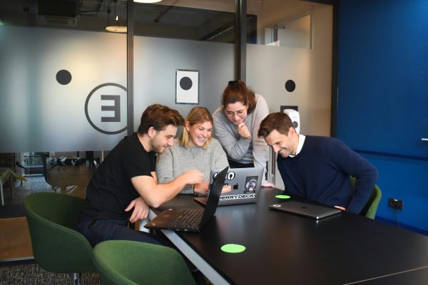 Quatre professionnels d'une trentaine d'années souriants, réunis autour d’un ordinateur dans un bureau moderne, illustrant un atelier MBTI à distance en entreprise.